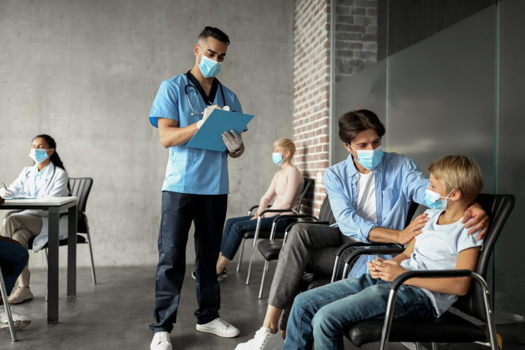 Friendly middle-eastern doctor talking to little boy and his father, filling medical chart. Diverse people in face masks sitting in hospital room or vaccination center waiting to get Covid-19 shot